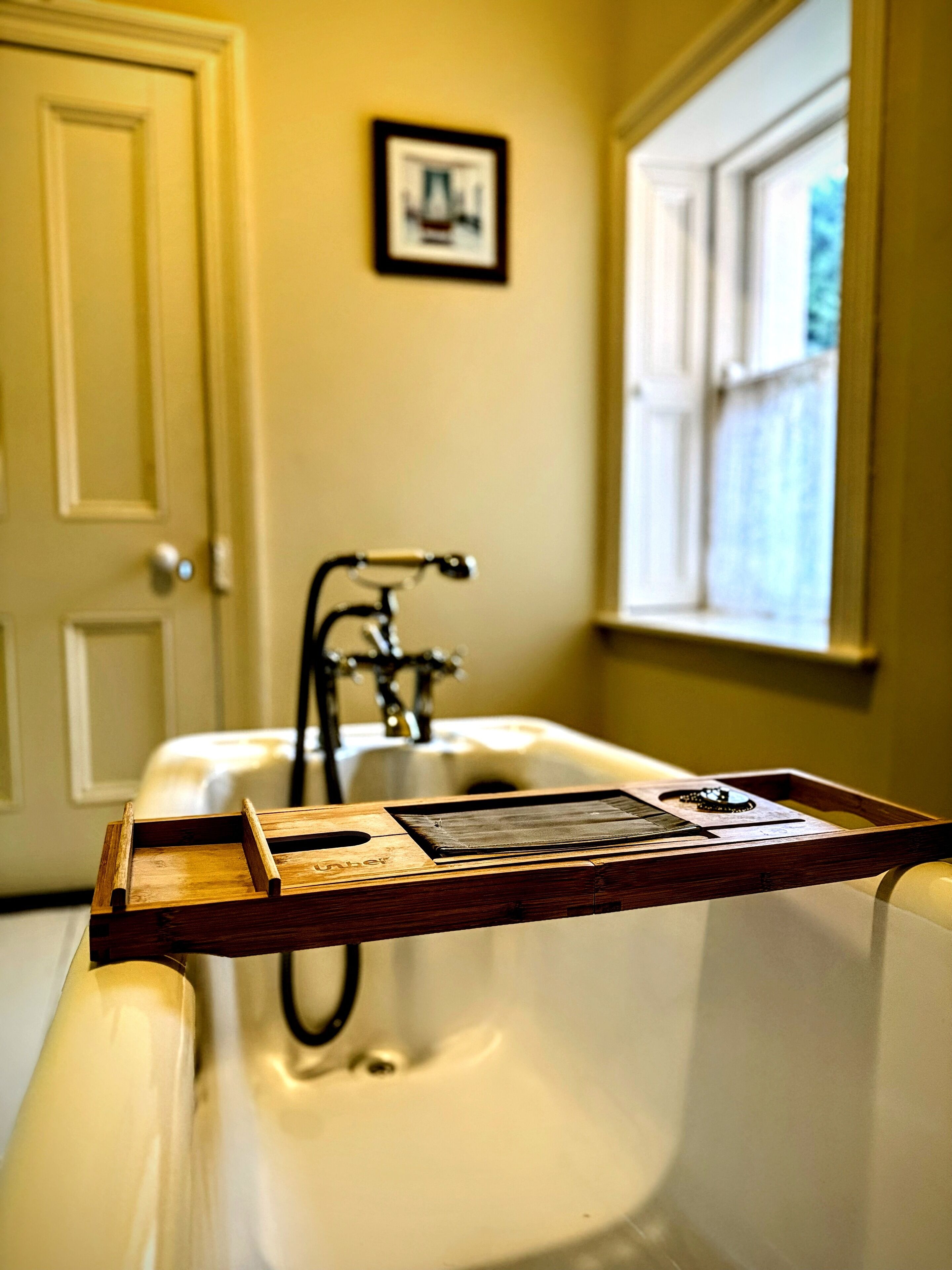 A bathroom with a wooden tray on the bathtub, a sink, a faucet, and a framed picture on the wall.