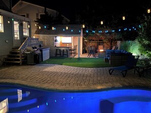Un jardin avec une piscine, des meubles de jardin, un espace barbecue et des guirlandes lumineuses.