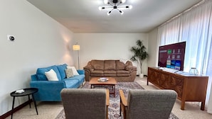 A living room with a blue sofa, a brown sofa, a wooden coffee table, two armchairs, a small side table, a floor lamp, a TV on a wooden cabinet, and a potted plant.