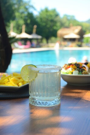 Et glas vand med et limeskive på et træbord, en salatskål og en swimmingpool i baggrunden.