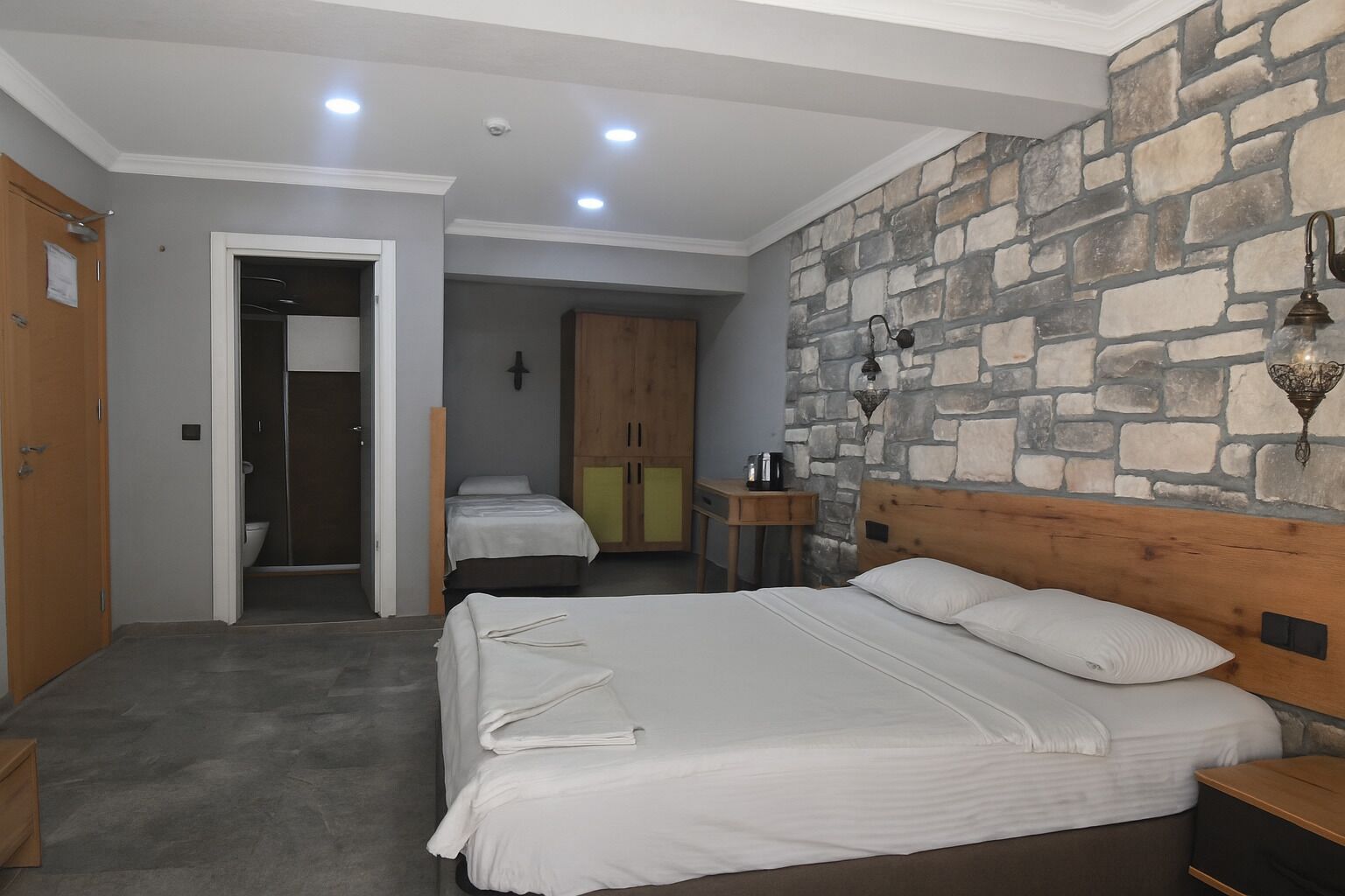 Une chambre avec un grand lit, une tête de lit en bois, un mur en pierre et une petite table avec une lampe.