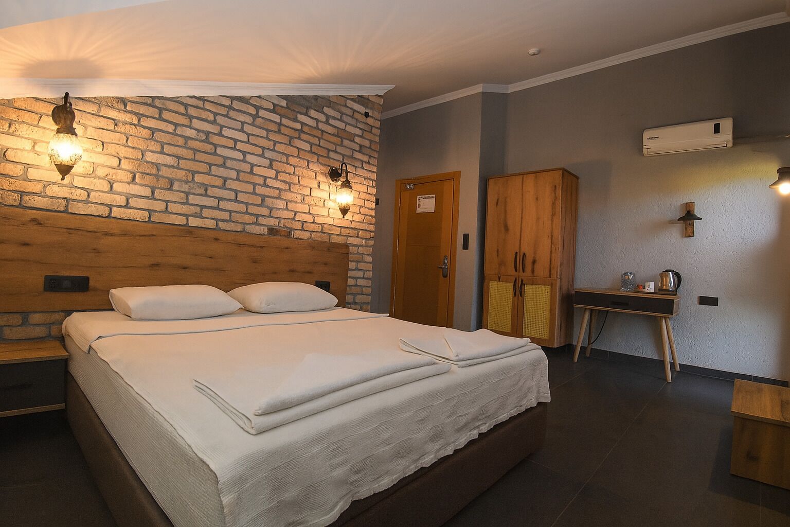 A hotel room with a brick-patterned headboard, a wooden wardrobe, a desk with a chair, and a bed with white linens.