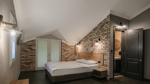 Un dormitorio con techo inclinado, paredes de ladrillo visto, cama con ropa blanca, mesita de noche de madera y una puerta oscura.