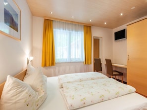 A hotel room with a bed, a desk, a chair, a TV, and a window with yellow curtains.