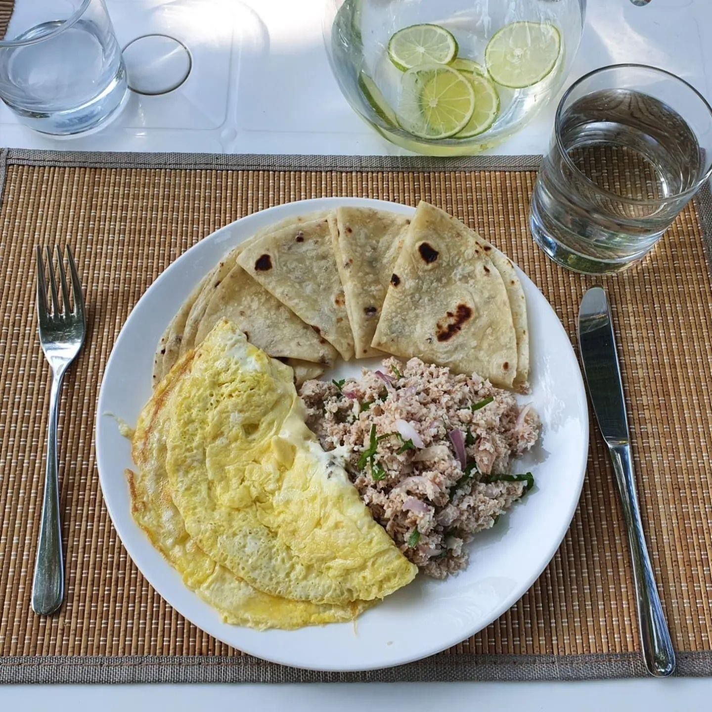 A plate with an omelette, flatbreads, and a meat dish, accompanied by a glass of water and a bowl of lime slices.