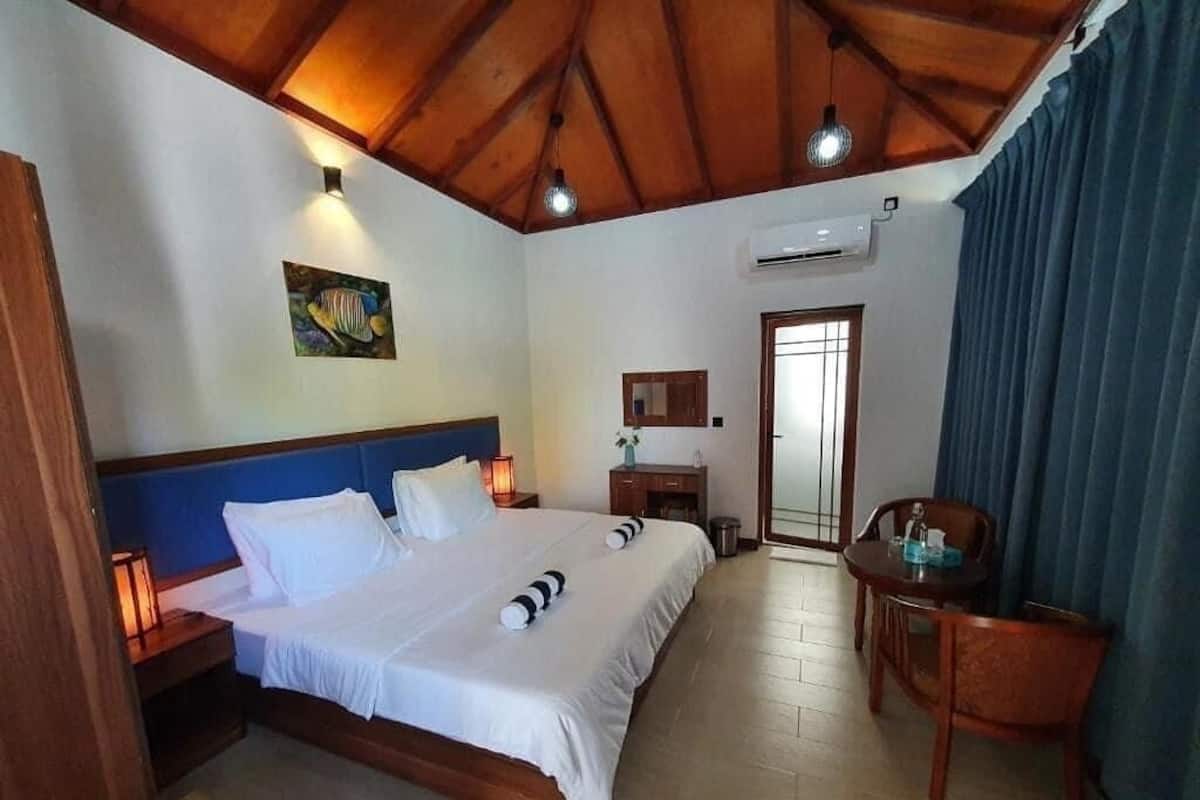 Habitación de hotel con techo de madera, cama con ropa blanca, escritorio con silla y ventana con cortinas.