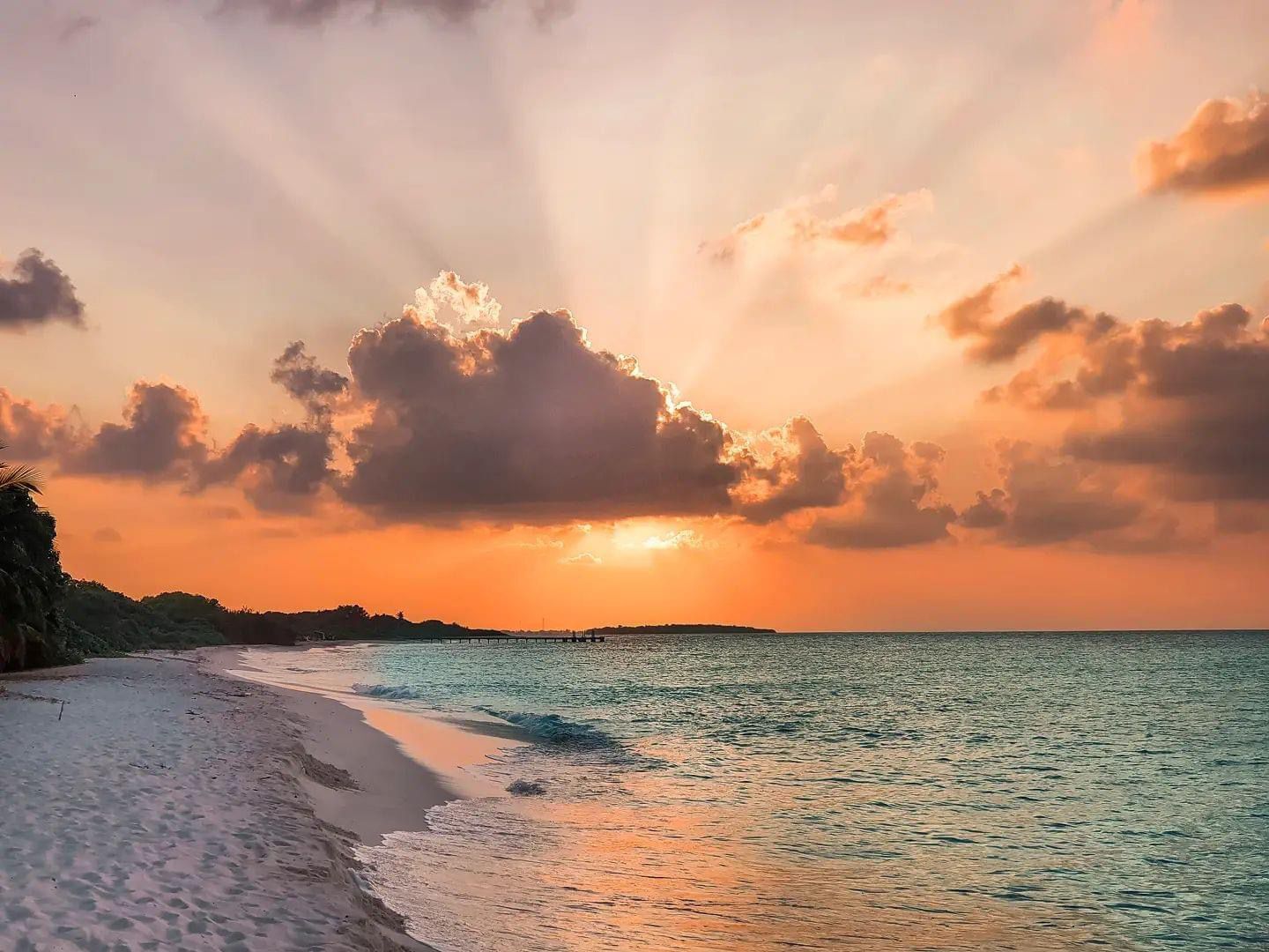 Una playa con un atardecer, mar tranquilo y un muelle a lo lejos.