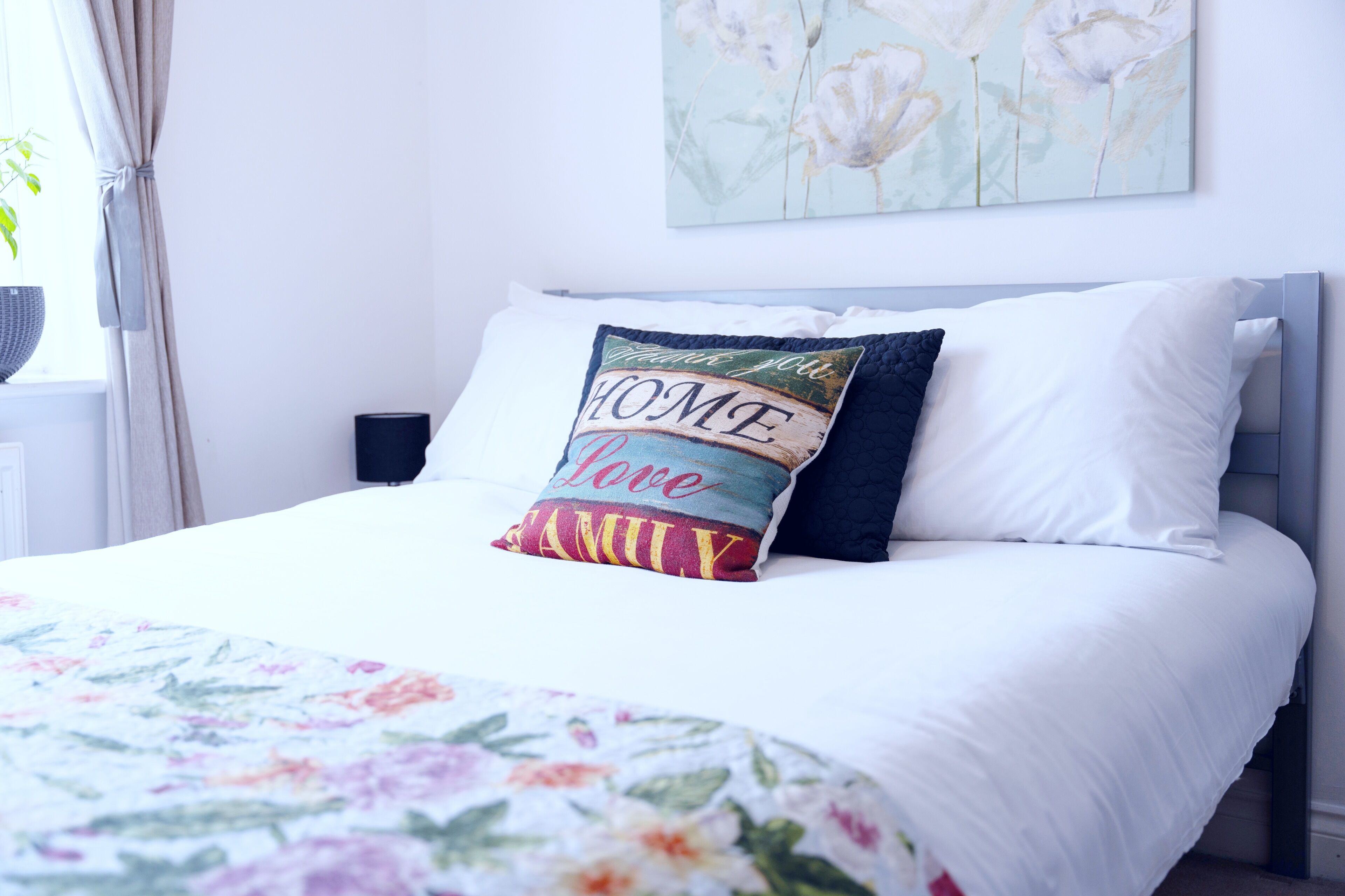 Een net opgemaakt bed met een bloemenbedekking, een kussen met de tekst 'HOME LOVE FAMILY' en een bloemen schilderij aan de muur.