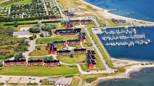 Une vue aérienne d’un quartier résidentiel situé sur la côte, avec de nombreux bâtiments aux toits rouges, une marina où sont amarrés de nombreux bateaux et une route menant à la plage.