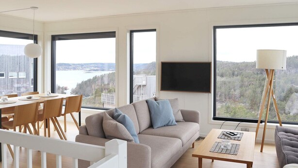 A modern living room with a sofa, dining table, and chairs. There is a large window with a view of a forest and a body of water. - 6 Person Holiday Home in Sondeled (Risør)