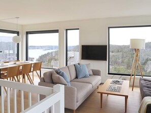 Un salon moderne avec un canapé, une table à manger et des chaises. Une grande fenêtre offre une vue sur une forêt et un plan d’eau.