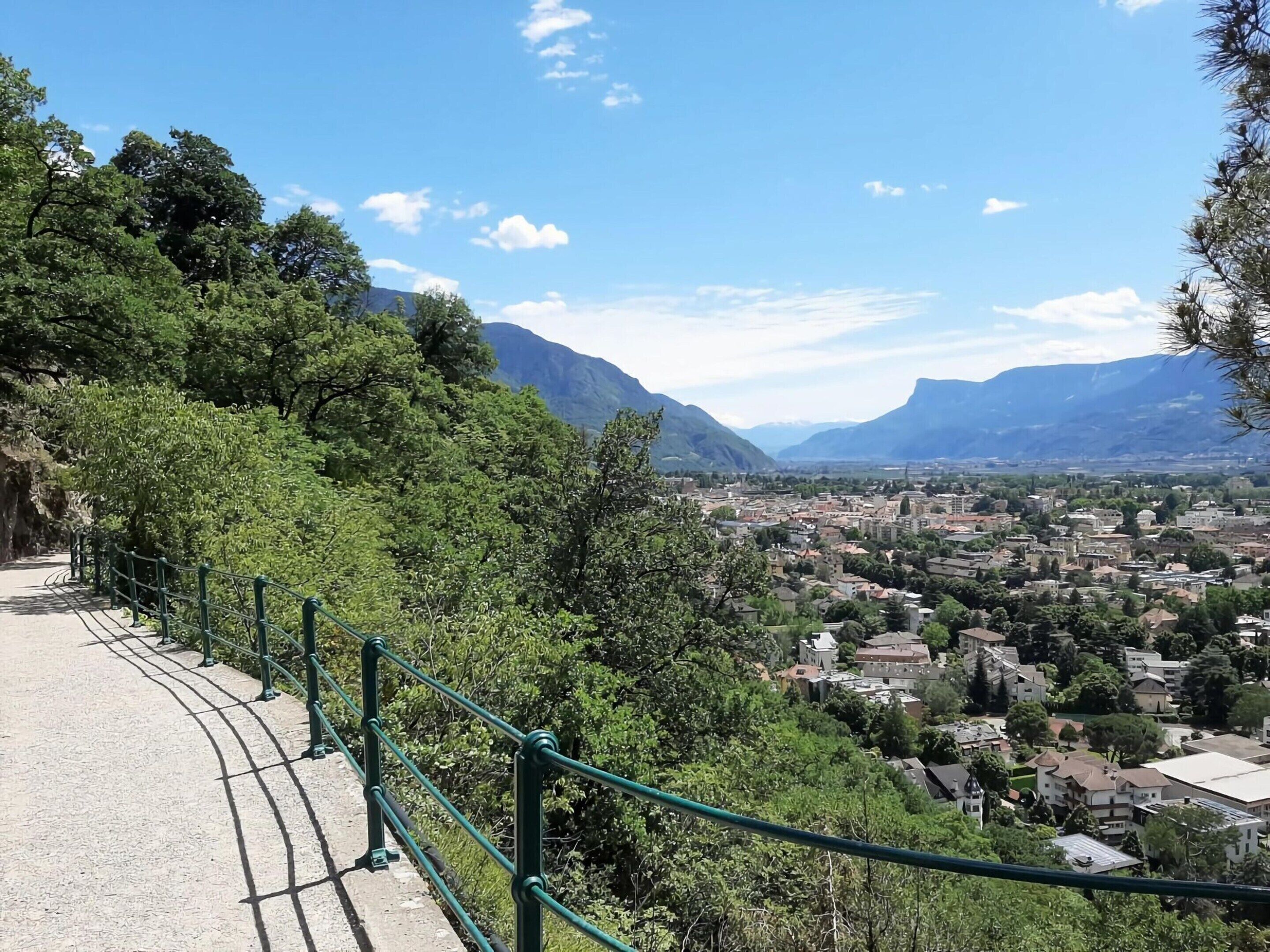 A scenic overlook with a metal railing, lush greenery, and a view of a town and mountains in the distance.