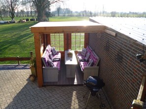 A covered outdoor seating area with a table and chairs, a barbecue grill, and a view of a grassy field. - Pleasant Apartment in Xanten (Xanten)