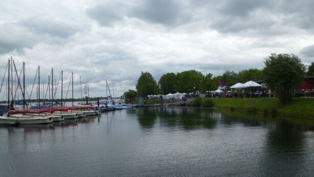 A calm lake with several boats docked on the left, a grassy area with trees and a building on the right, and a cloudy sky above. - Pleasant Apartment in Xanten (Xanten)
