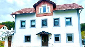 A two-story building with a white facade and red-tiled roof. It has multiple windows, a central entrance with a dark door, and a small porch area. - Rustic holiday home for 12 people (Warmensteinach)