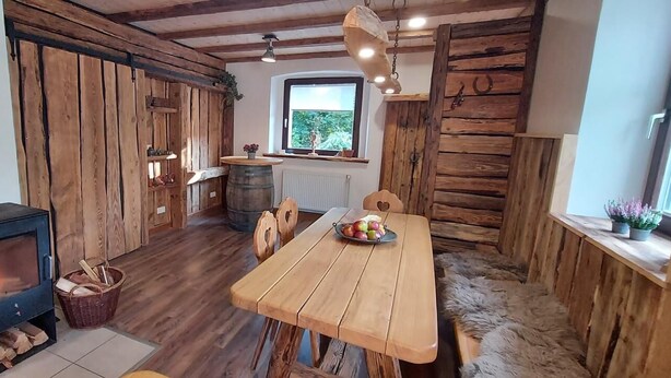 A rustic interior with a wood-burning stove, wooden walls, a dining table with fruit, and a window with a view. - Rustic holiday home for 12 people (Warmensteinach)