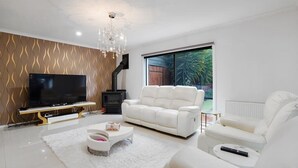 Una sala de estar moderna con un televisor de pantalla plana, sofás de cuero blanco, una araña y un amplio ventanal con vistas a la vegetación.