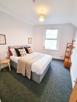 A bedroom with a bed, a bedside table, a chair, a window, and a wall-mounted shelf.