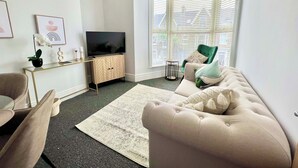 Olohuoneessa on sohva, televisio puisen kaapin päällä, pieni pöytä ja ikkuna, jossa on sälekaihtimet.