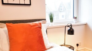 Siististi pedattu sänky, jossa on valkoisia ja oransseja tyynyjä, musta lamppu yöpöydällä ja ikkuna, josta avautuu näkymä rakennuksiin.