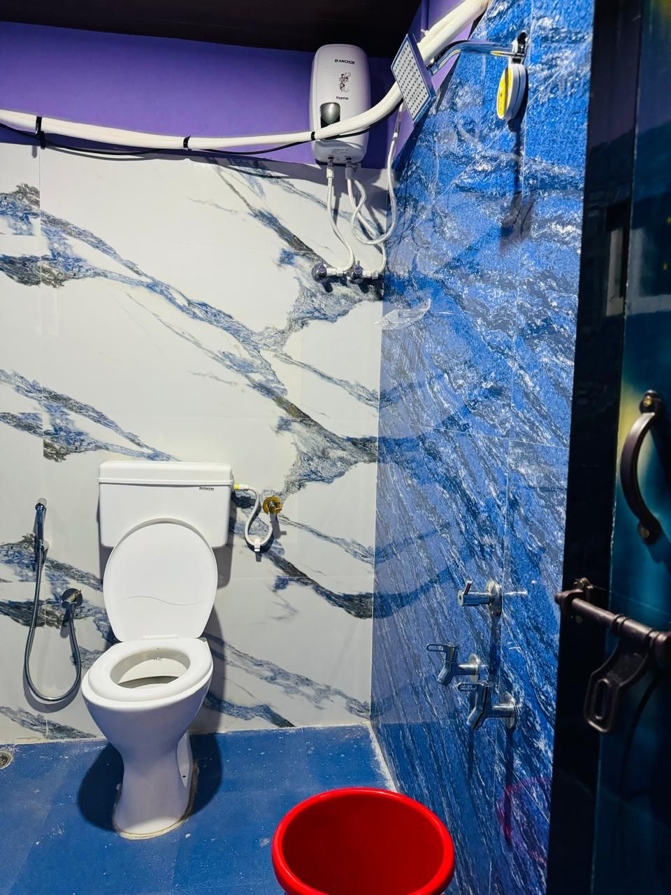 A bathroom with a white toilet, a red bucket, a hand dryer, and a soap dispenser on a purple wall.