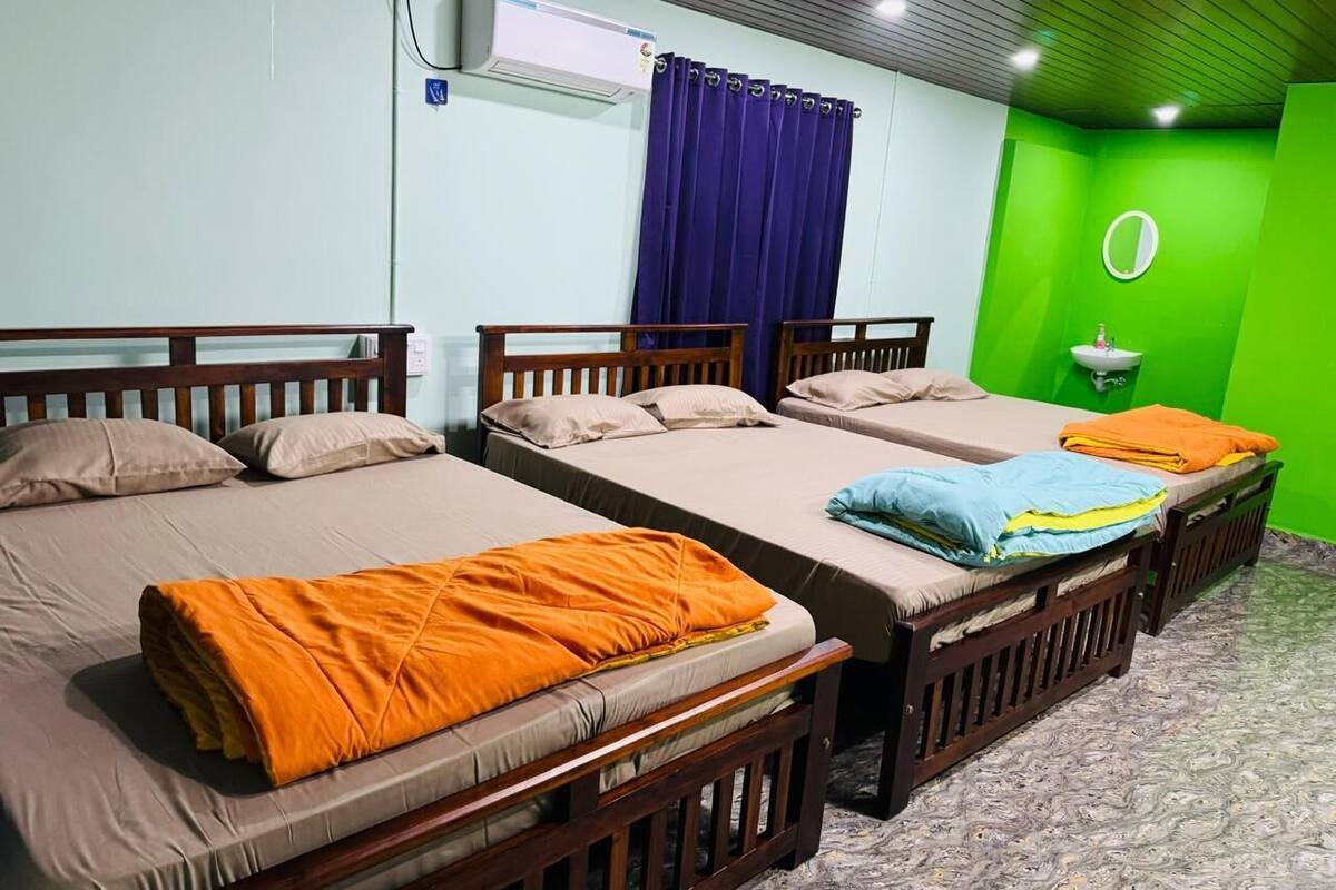 Ein Hotelzimmer mit vier Einzelbetten, einer grünen Akzentwand, einem runden Spiegel und einer Klimaanlage.