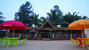 Een restaurant aan het strand met terras, palmbomen en kleurrijke hekken.