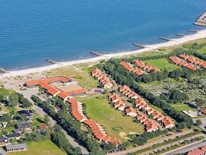 An aerial view of a coastal resort with multiple bungalow-style buildings, a sandy beach, and a rocky breakwater.