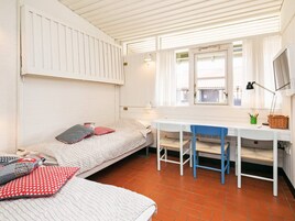Una habitación pequeña y funcional con una cama individual, un escritorio y una silla. Cuenta con una ventana con cortinas, una lámpara en el escritorio y una mesita con un cesto y bolígrafos.