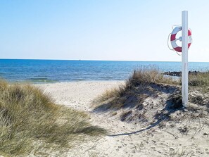 A beach with a lifebuoy attached to a pole, clear blue water, and sandy dunes with grass.