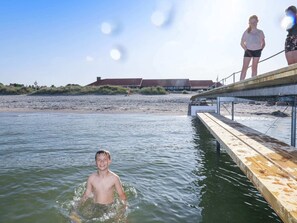 Une jetée en bois s’étendant dans une étendue d’eau, avec une plage de sable et un bâtiment visible au loin.