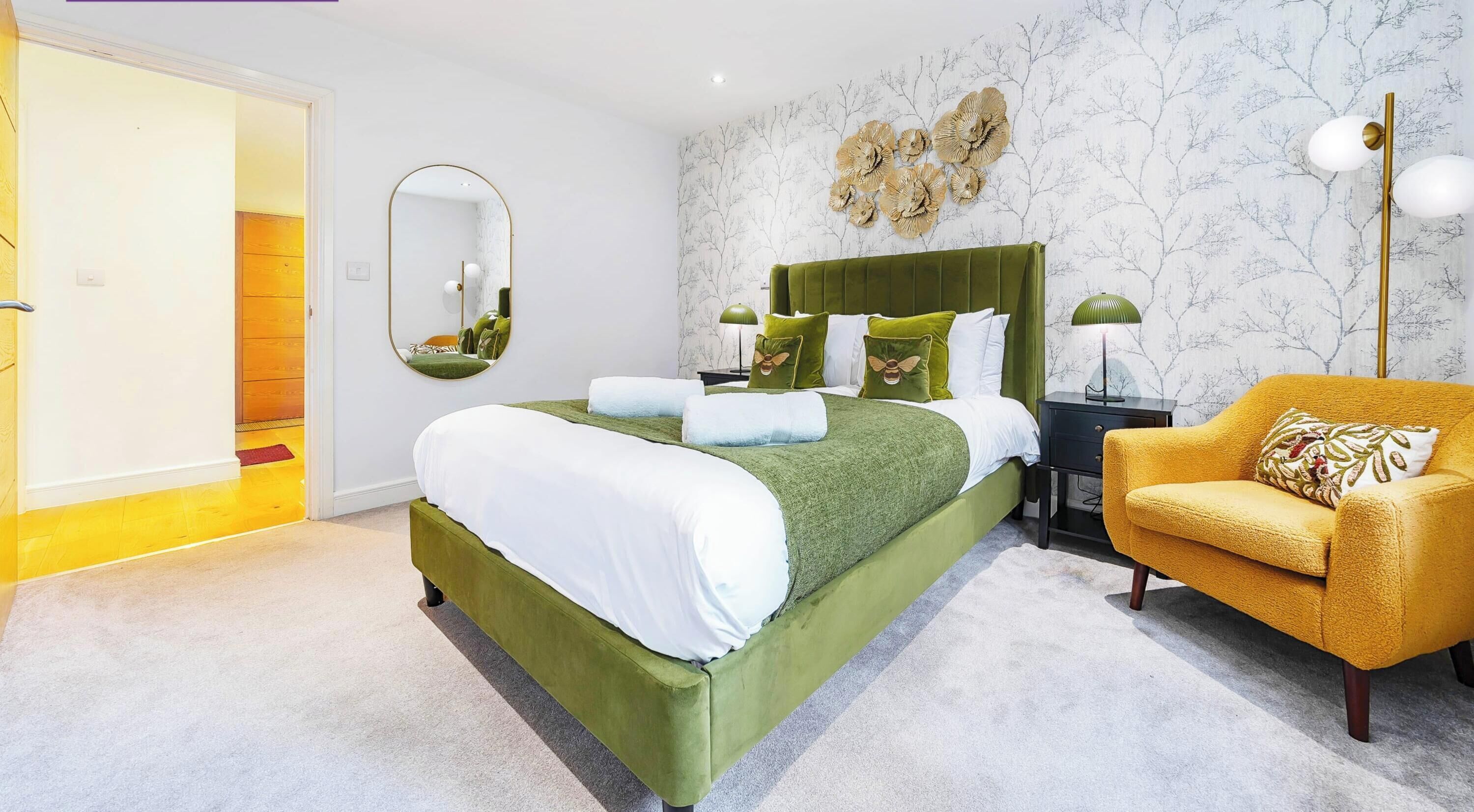 Une chambre avec un lit vert, un fauteuil jaune, un miroir et un mur orné d’un motif représentant un arbre.