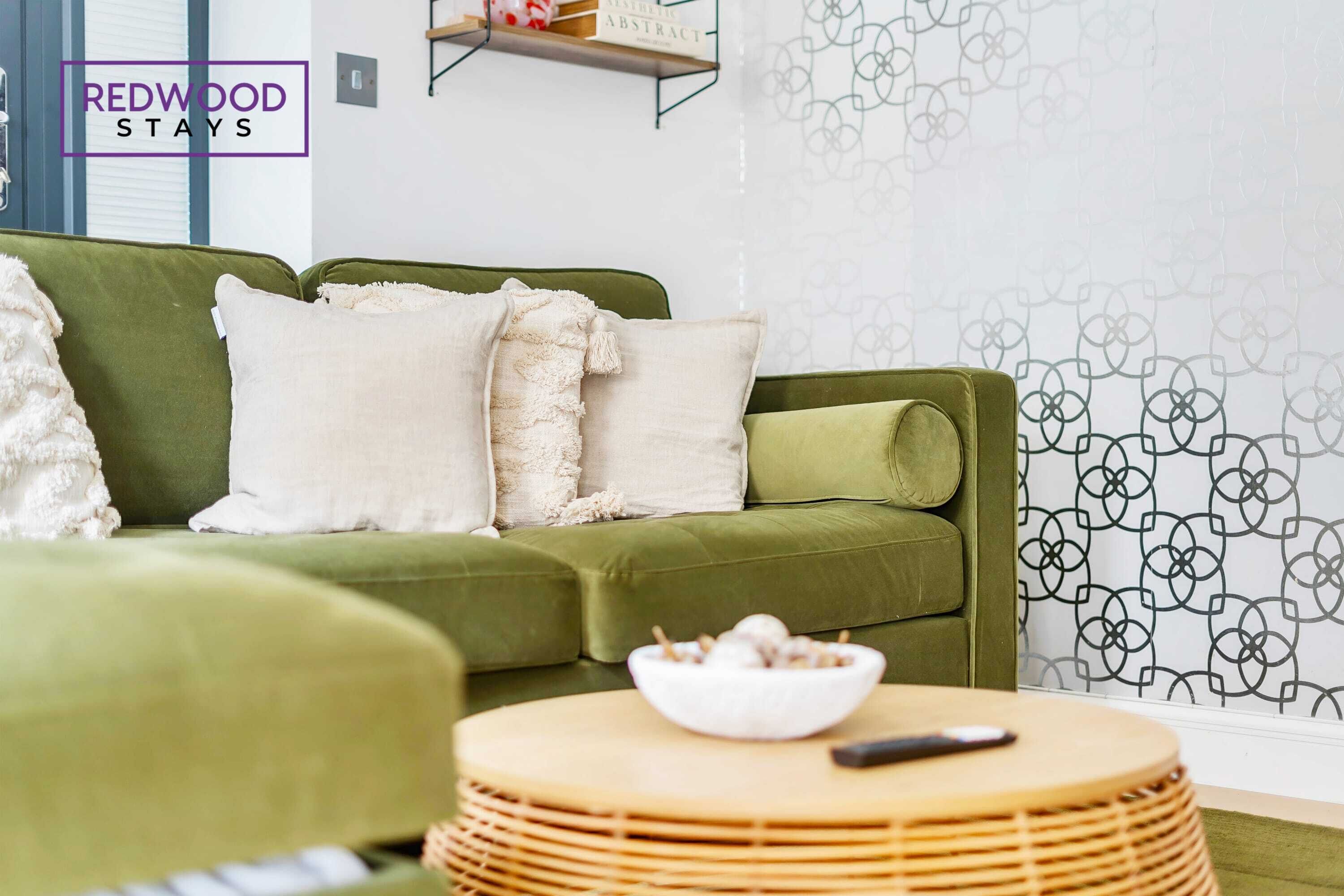 A green sofa with white cushions, a wicker side table with a bowl and remote, and a patterned wall.