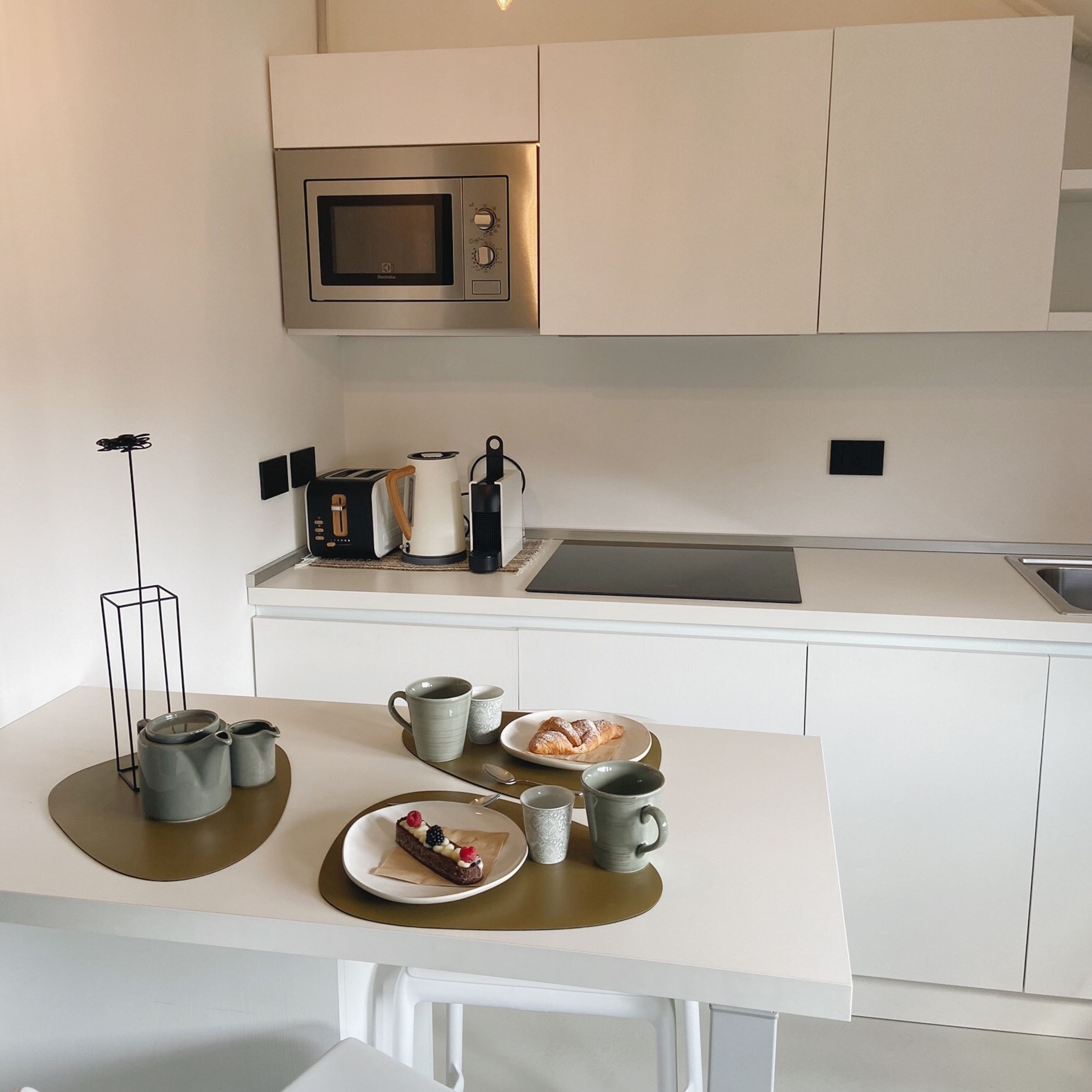 A modern kitchen with white cabinetry, a stainless steel microwave, a coffee maker, and a breakfast setting with croissants and a dessert on the counter.