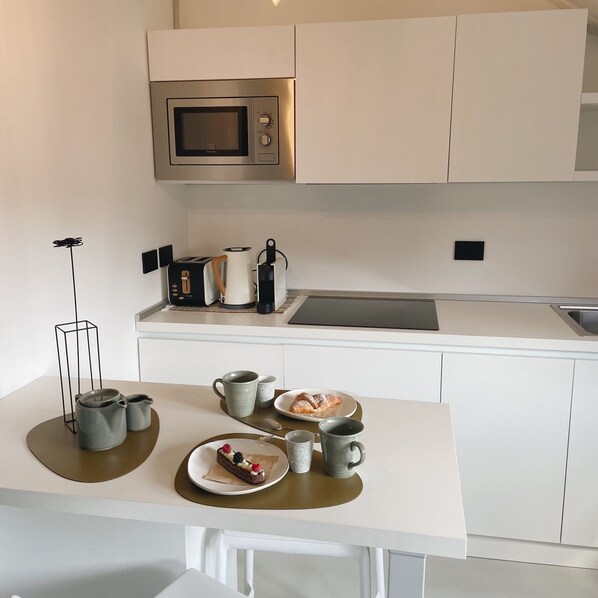 Una cocina moderna con armarios blancos, un microondas de acero inoxidable, una cafetera y una mesa para desayunar con cruasanes y un postre en la encimera.