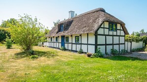 Une maison traditionnelle à colombages, avec un toit de chaume, entourée d’un champ herbeux et d’arbres.