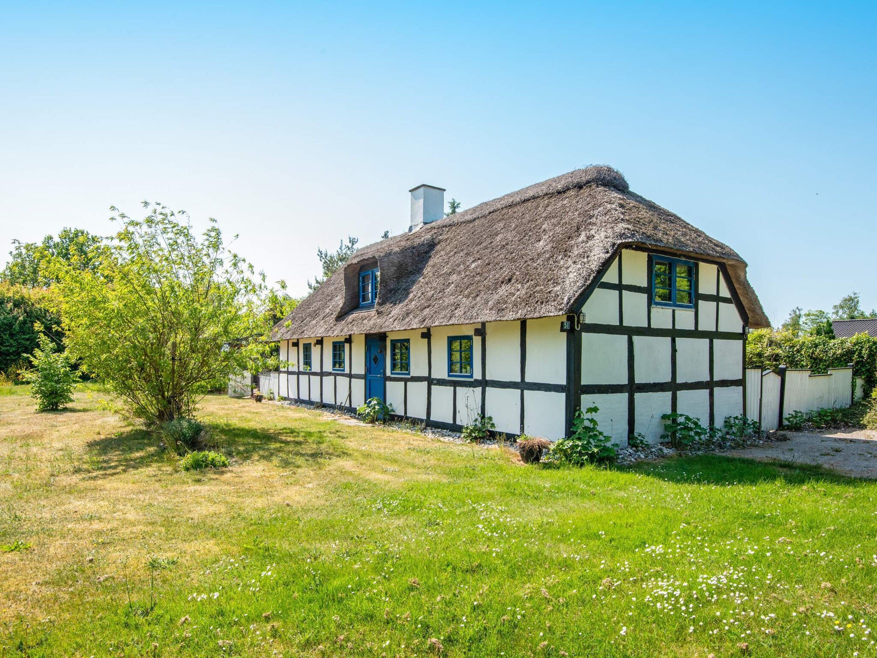 A traditional half-timbered house with a thatched roof, surrounded by a grassy field and trees.