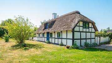 A traditional half-timbered house with a thatched roof, surrounded by a grassy field and trees.