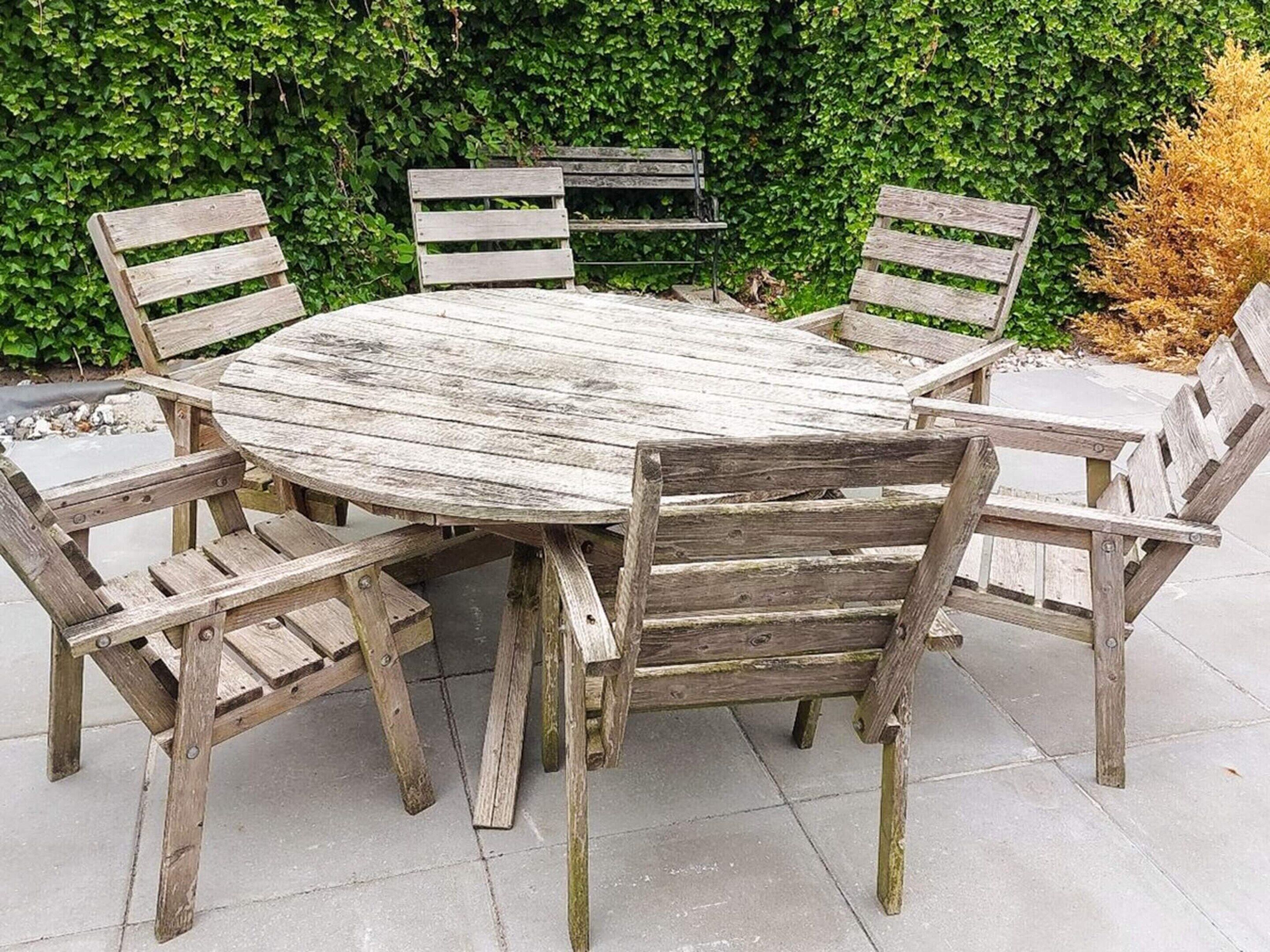 A round wooden table with six chairs, set outdoors with a hedge in the background.