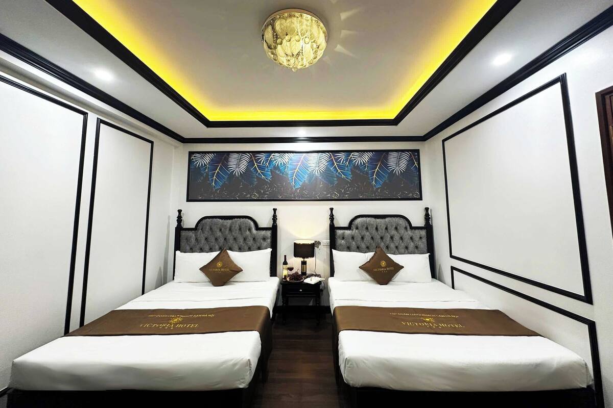 A hotel room with two beds, a chandelier, a large leaf-patterned artwork, and a brown bedspread.