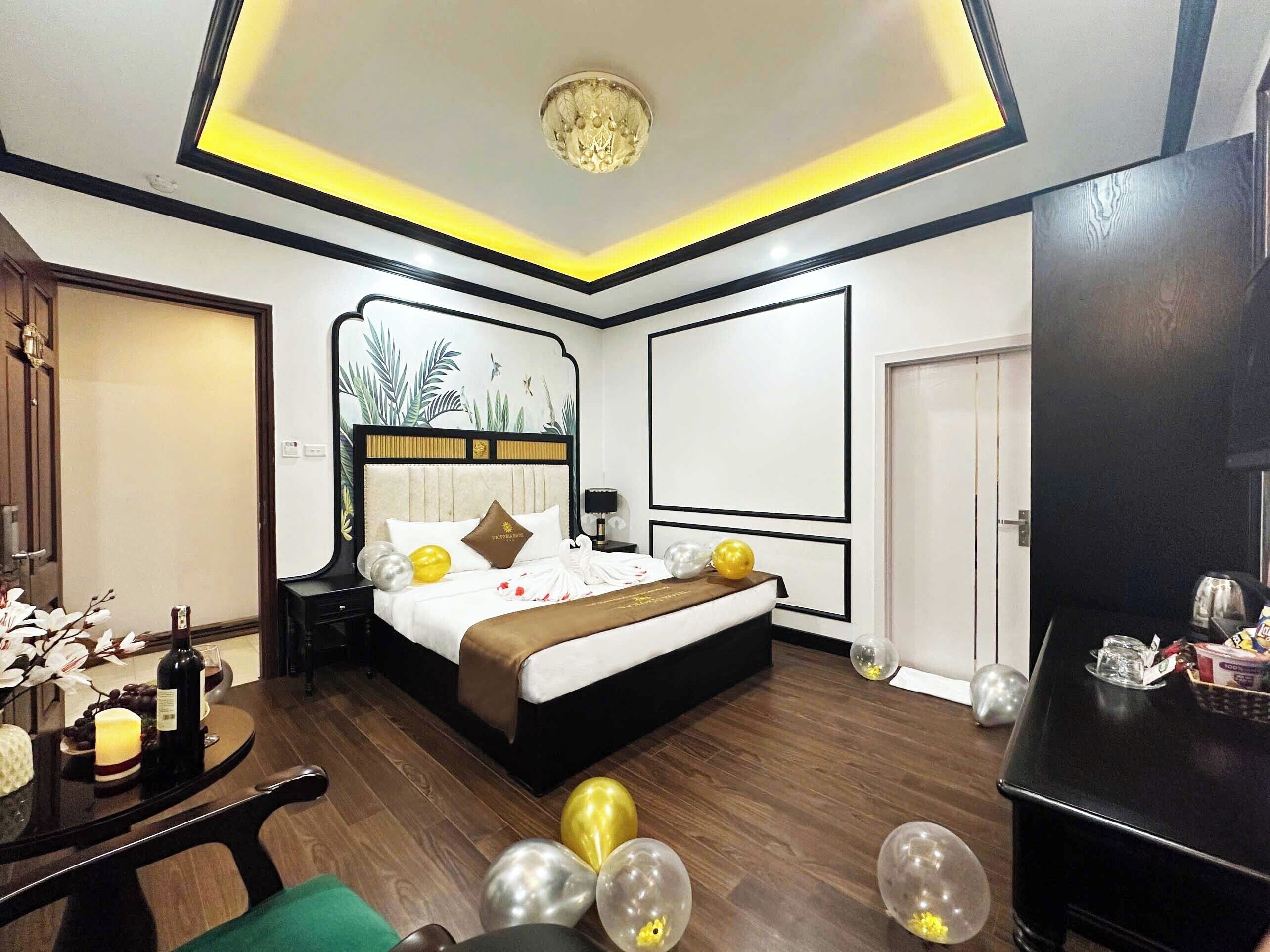 A modern hotel room with a large bed, a desk with a chair, a chandelier, and a wall with a tropical mural.