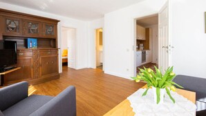 A modern apartment with a wooden dining table, two grey sofas, a kitchen, and a living area with a TV. - Pleasant Apartment in Suddorf (Nebel)