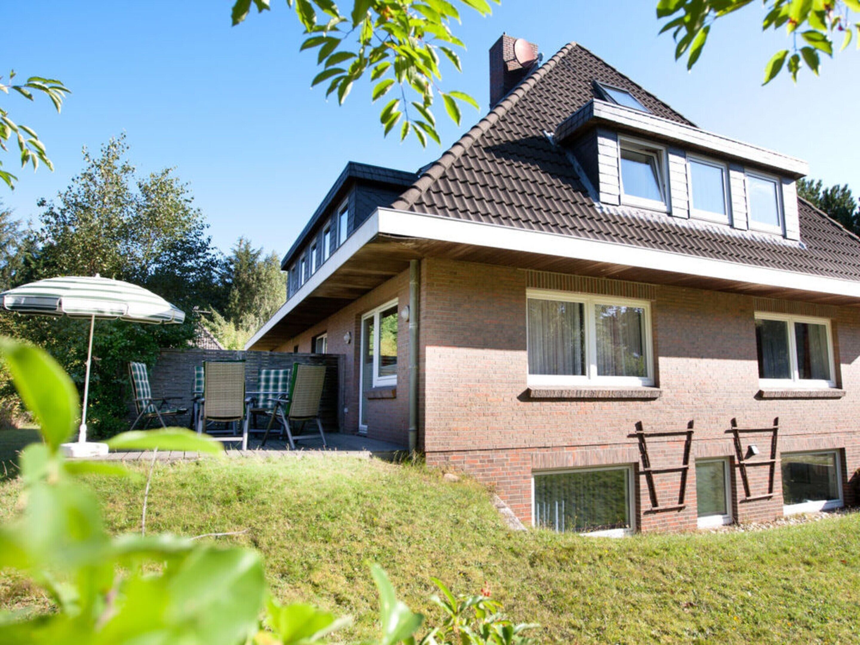 Uma casa de dois andares com fachada de tijolos, telhado inclinado e chaminé visível. Possui um pátio com mesa e cadeiras, além de um grande guarda-sol.