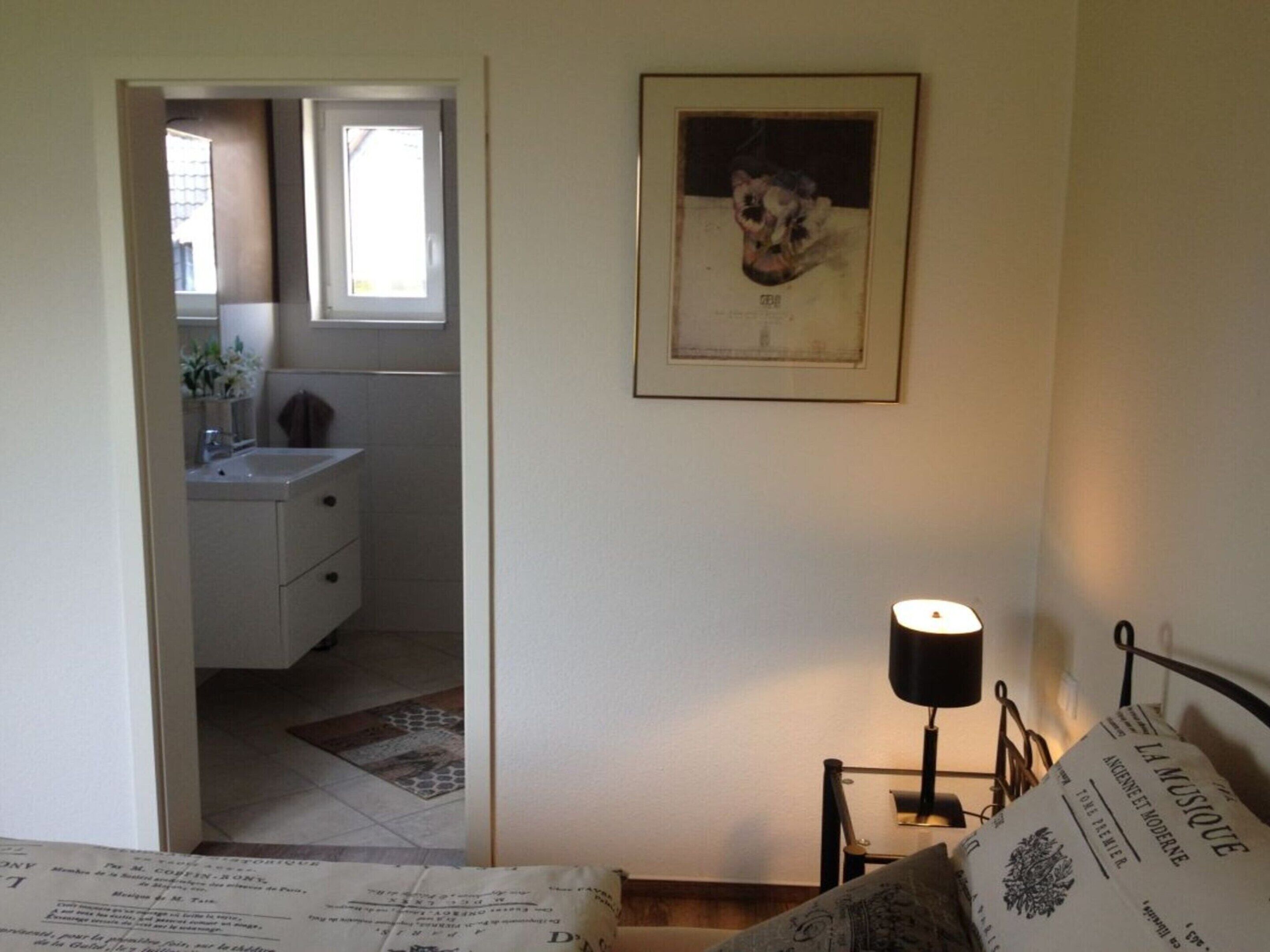 A bedroom with a bed, a bedside table with a lamp, a framed picture on the wall, and a view into the bathroom.