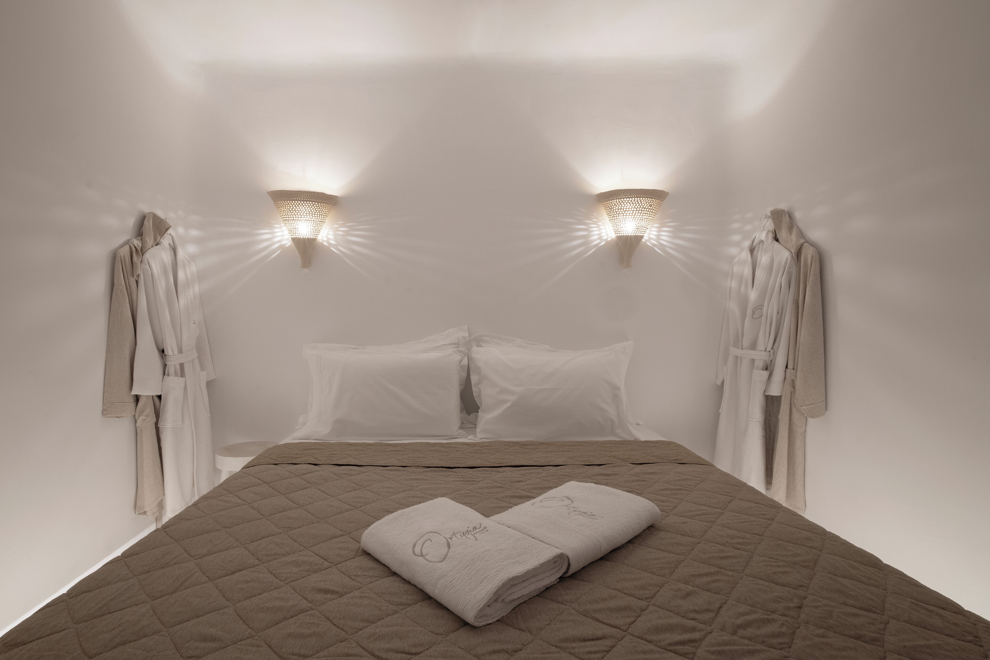 A neatly made bed with a quilted cover, two towels with text, and two wall-mounted light fixtures.