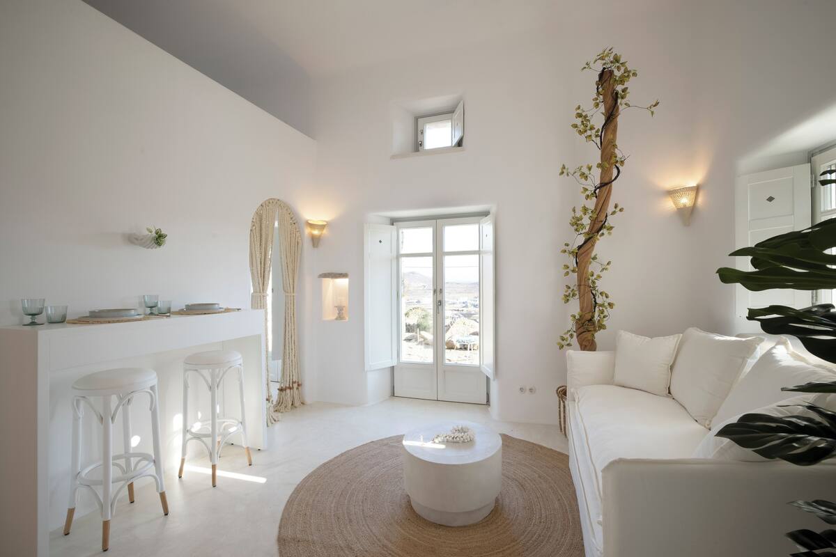 A bright, white interior with a round coffee table, white bar stools, a white sofa, and a large plant.