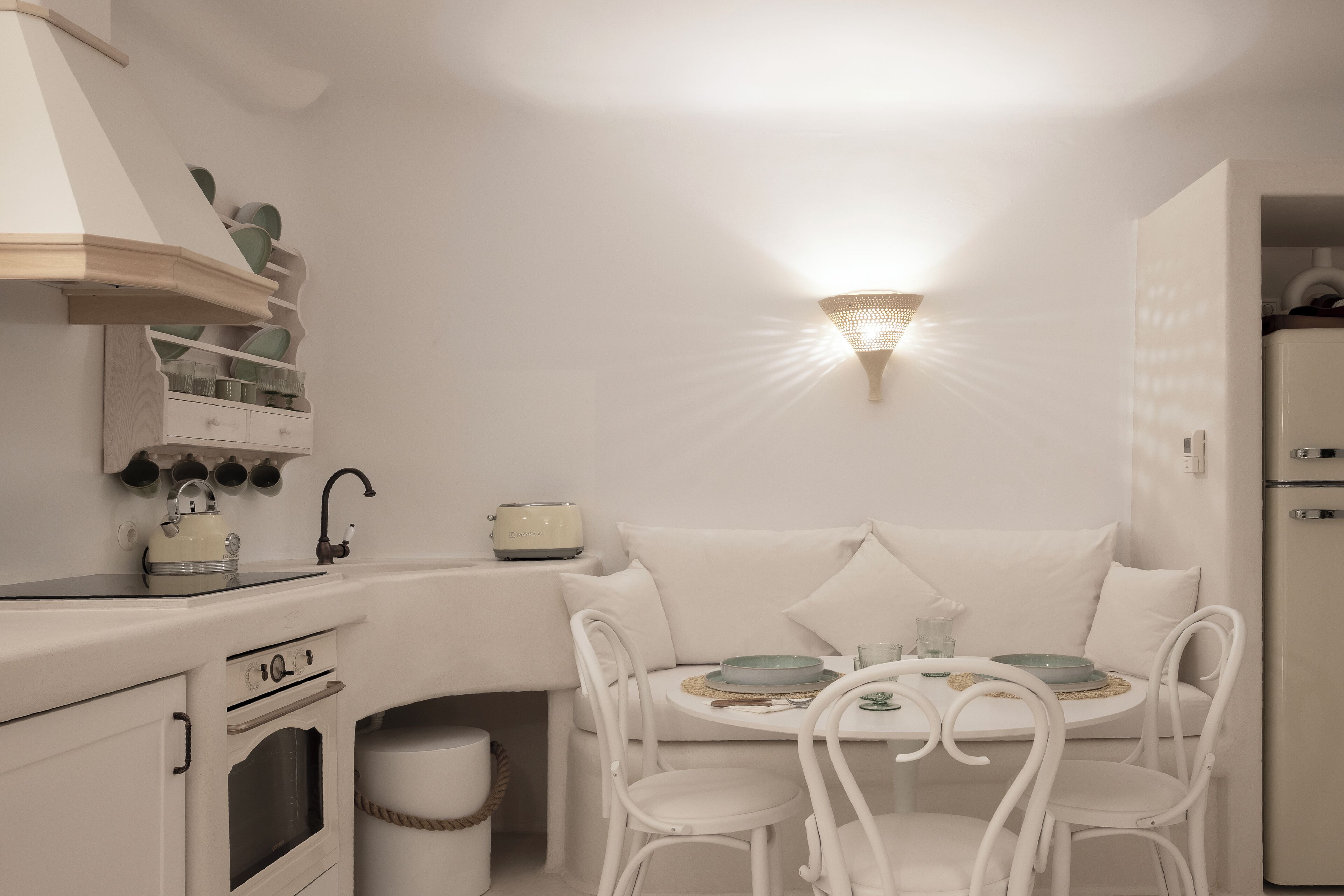 A compact kitchen with white appliances, a dining area with a round table and chairs, and a wall-mounted light fixture.