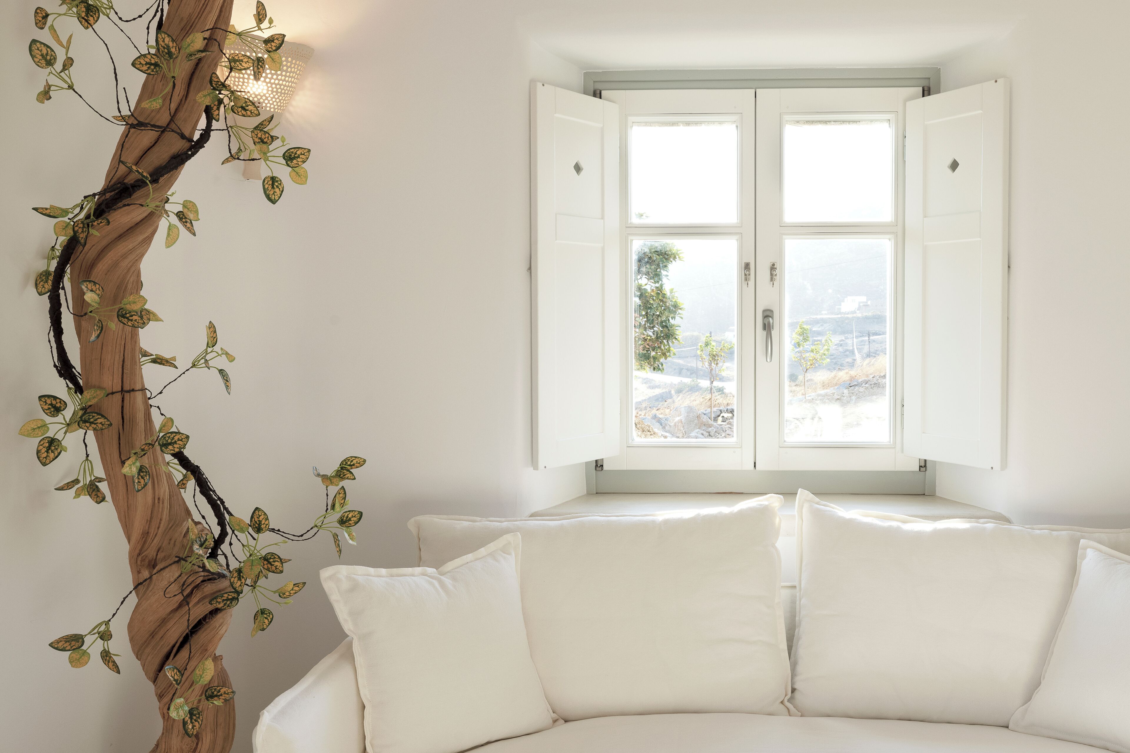 Una habitación con un sofá blanco, una escultura de árbol de madera y una ventana con contraventanas abiertas.