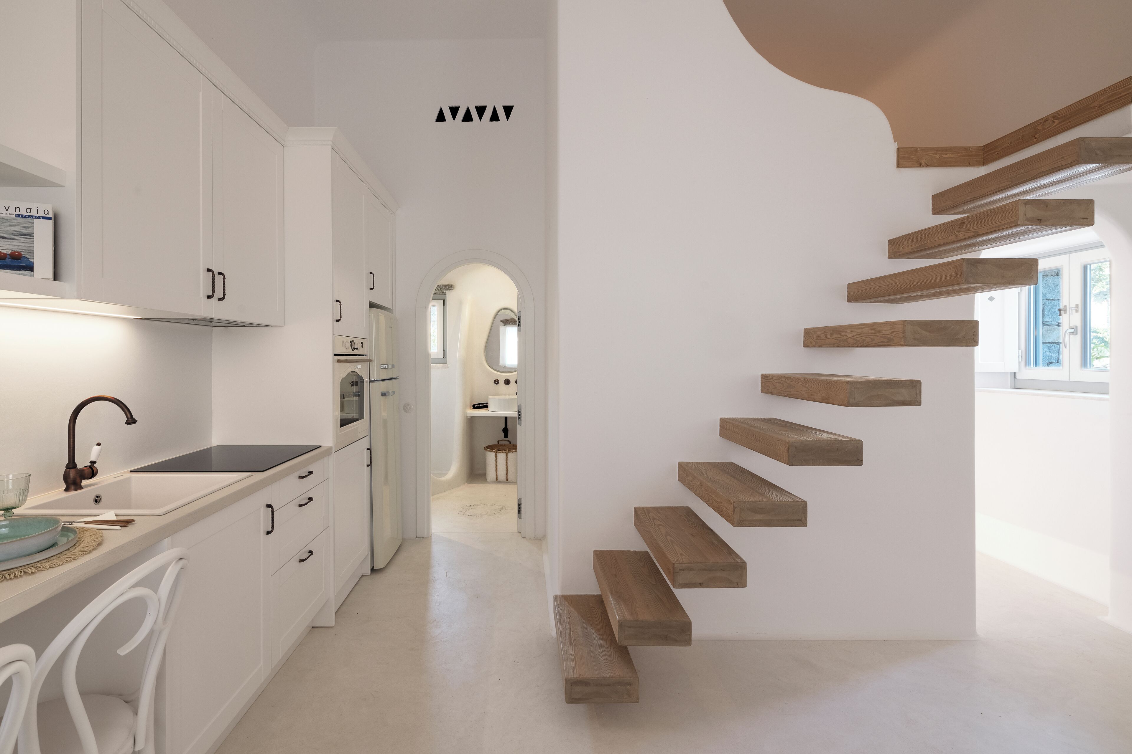 Una cocina moderna con armarios blancos, una escalera de madera y vista al comedor.