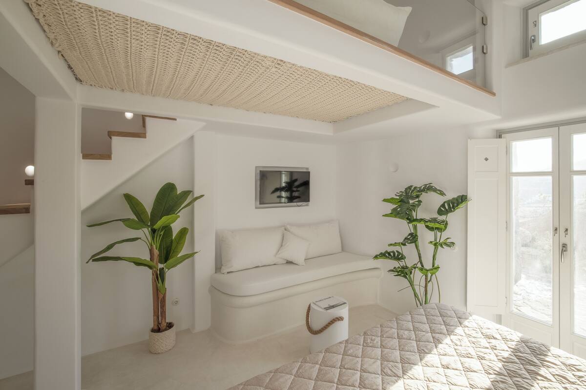 A modern, minimalist room with a white sofa, a large window, and a staircase leading to an upper level.
